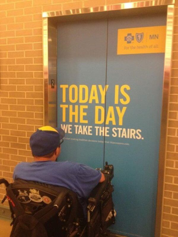 Wheelchair user in front of a shut elevator with the inscription “Today is the day we take the stairs”