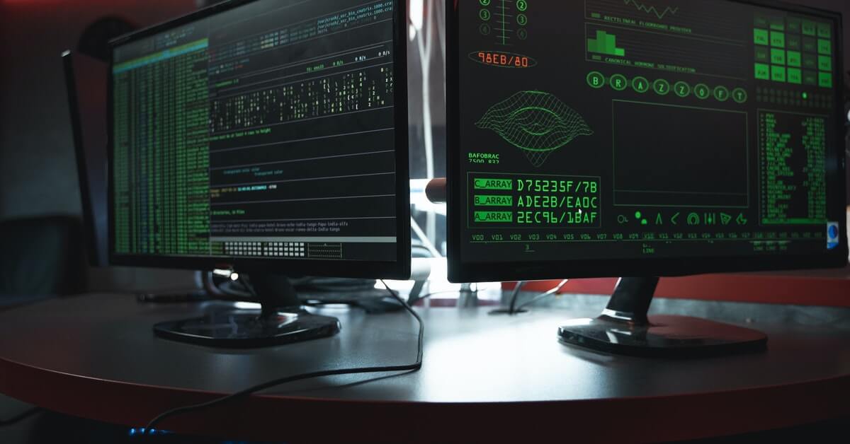 Two monitors placed side-by-side on a table displaying strings of codes in a dark-view mode.